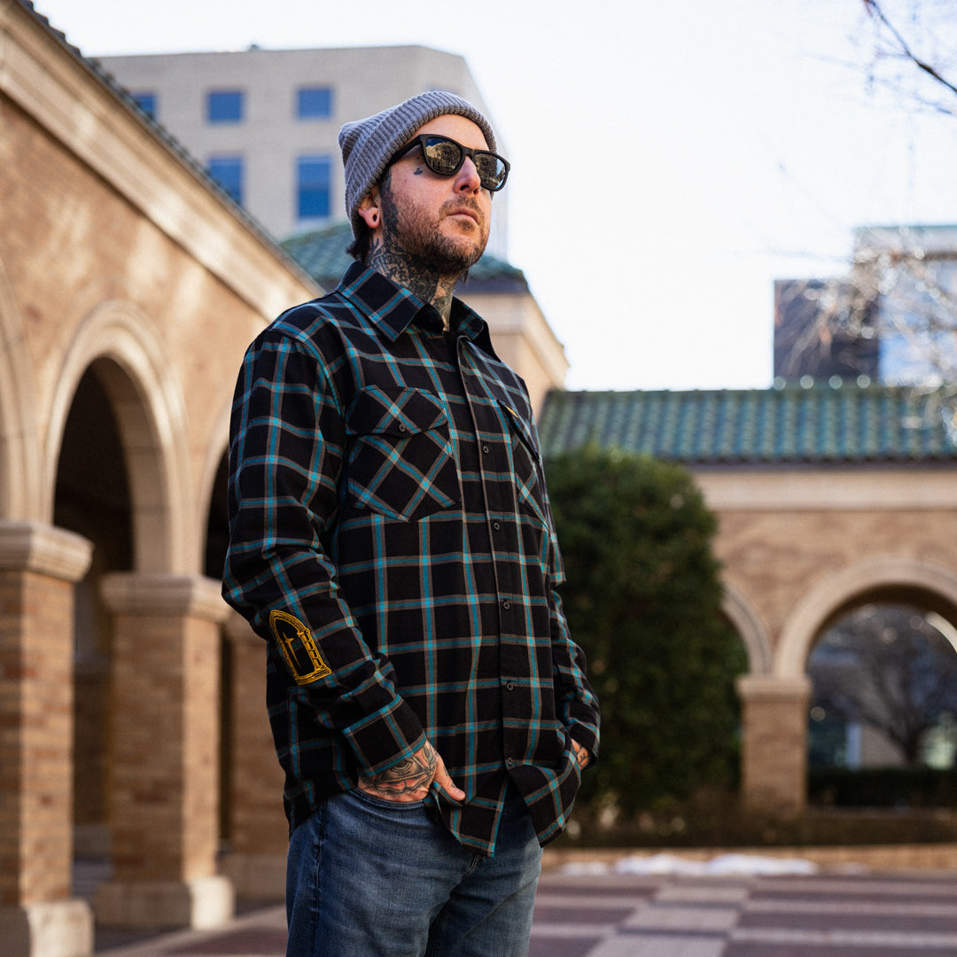 Man wearing a plaid shirt and sunglasses standing in front of architectural structures.
