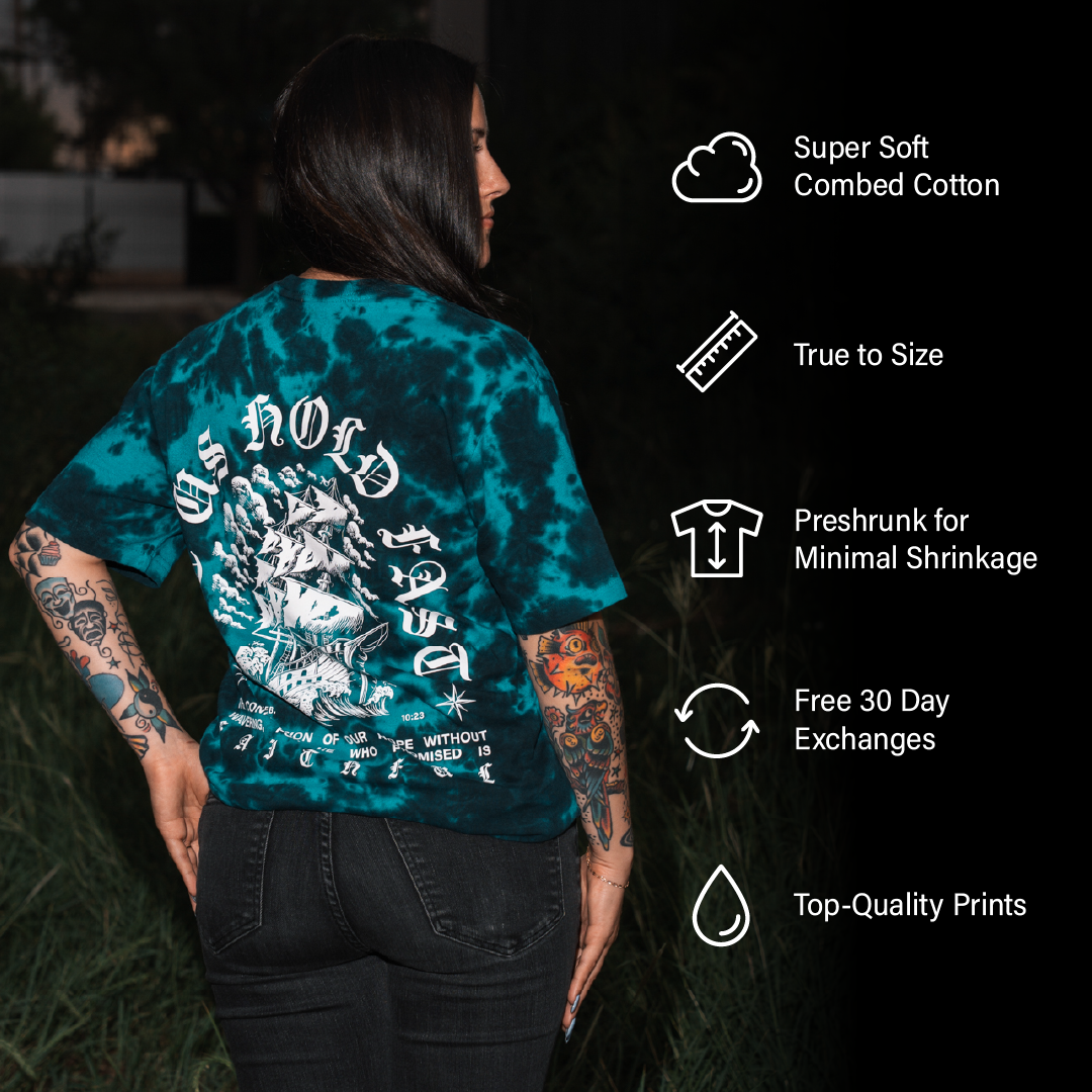 A woman with long dark hair and tattoos stands outdoors at night wearing a teal and black tie-dye t-shirt with a Christian nautical graphic on the back based on Hebrews 10:23.