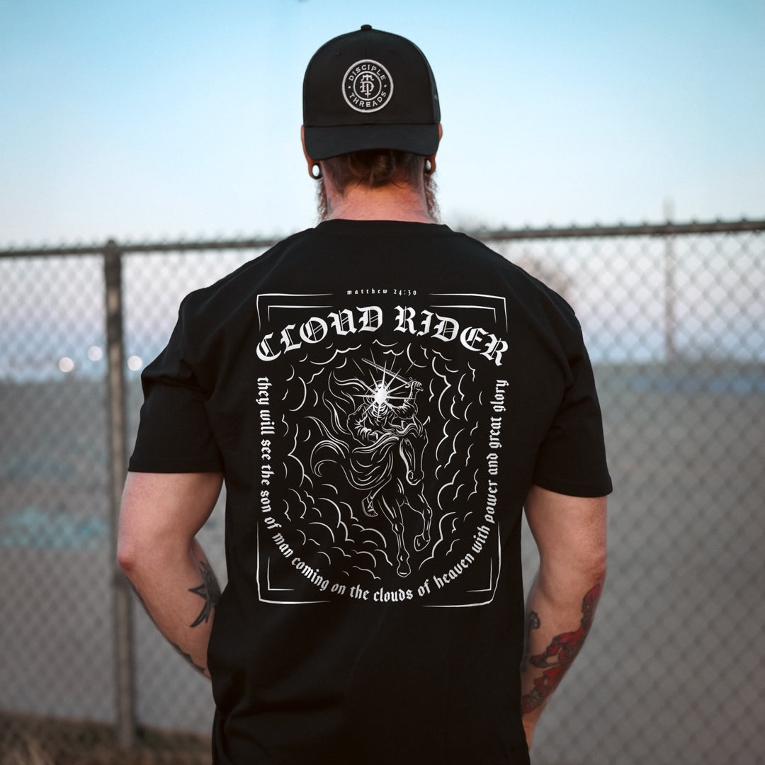 Person wearing a black t-shirt with 'Cloud Rider' design and cap, standing against a chain-link fence.