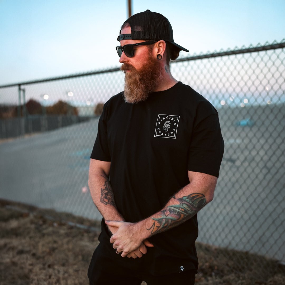 Man with tattoos wearing a black t-shirt with a logo, sunglasses, and a cap, standing in front of a chain-link fence.