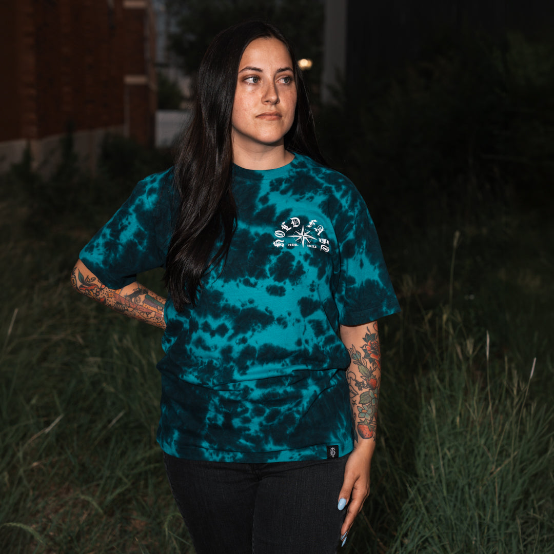 A woman with long dark hair and tattoos stands outdoors at night wearing a teal and black tie-dye t-shirt with a white graphic on the chest, paired with black pants.