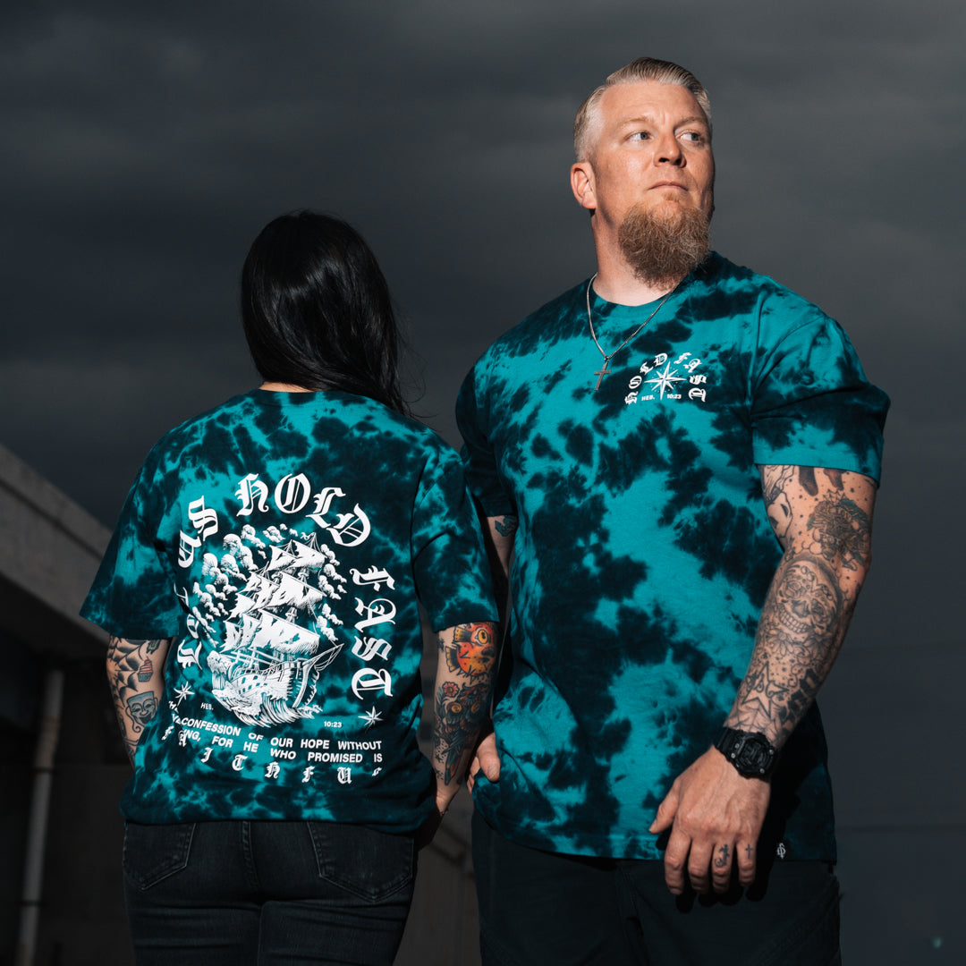 Two models wearing matching teal and black tie-dye t-shirts with white graphic prints, photographed outdoors against a dark cloudy sky.