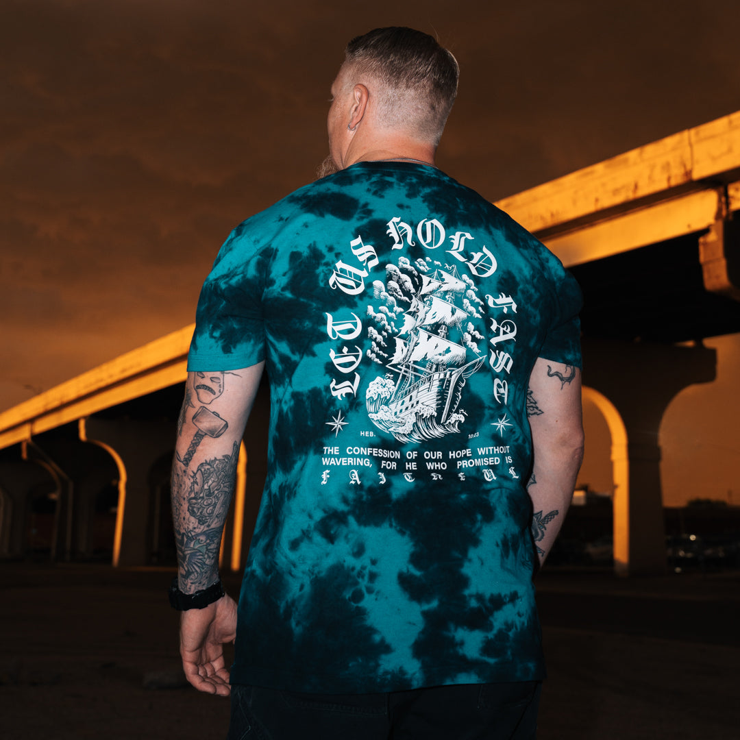 A man viewed from behind wearing a teal and black tie-dye t-shirt with a large Hebrews 10:23 graphic on the back, standing in front of an illuminated concrete overpass at night.