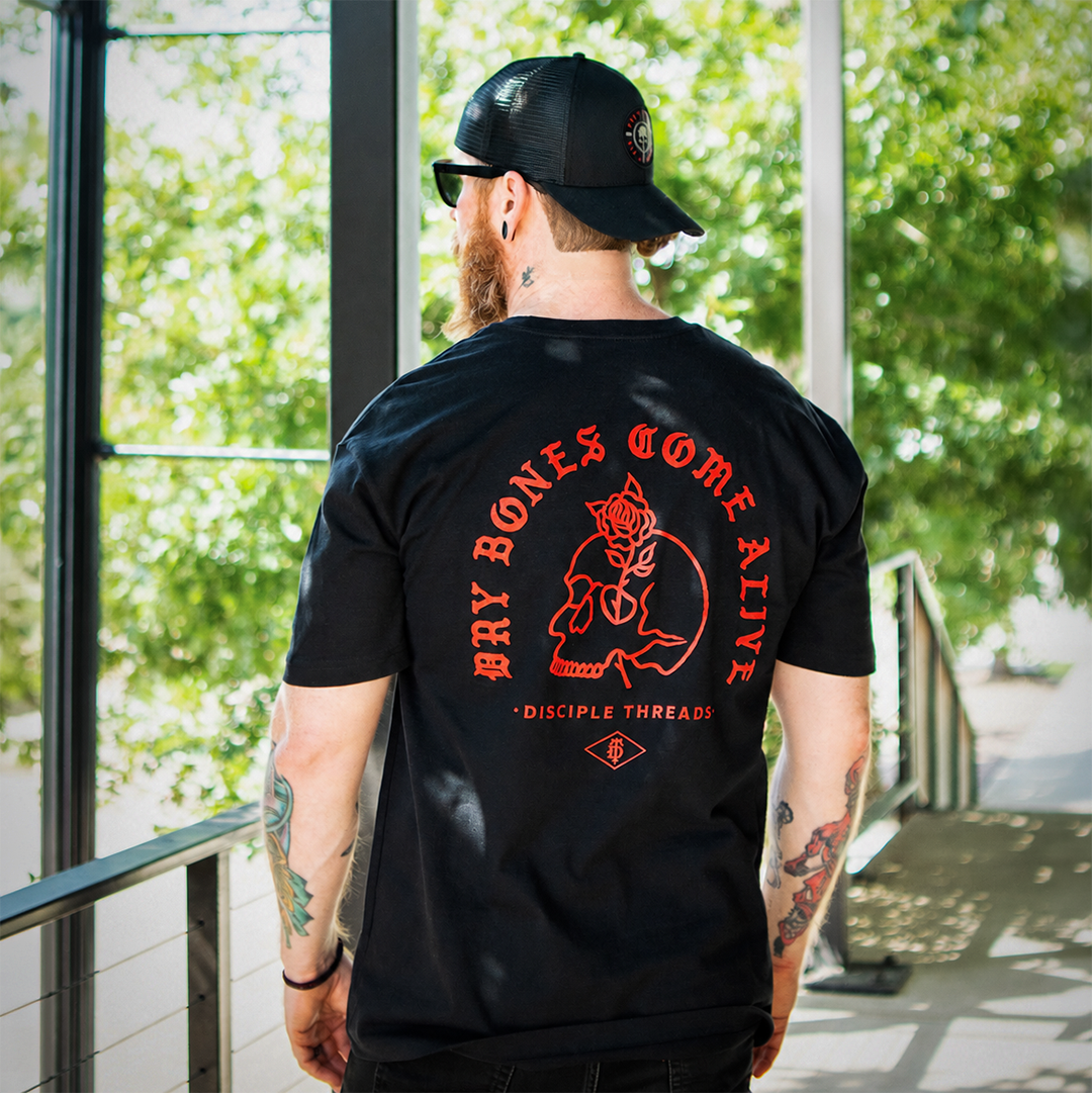 A man viewed from behind wearing a black t-shirt with a large red graphic on the back that reads 'DRY BONES COME ALIVE' and 'DISCIPLE THREADS'. He is also wearing a black trucker hat and standing on a balcony with green trees in the background.