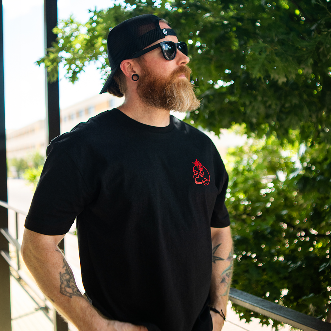 A man with a red beard, sunglasses, and a black cap wearing a black t-shirt with a small red graphic on the left chest, standing outdoors on a balcony with green foliage in the background.