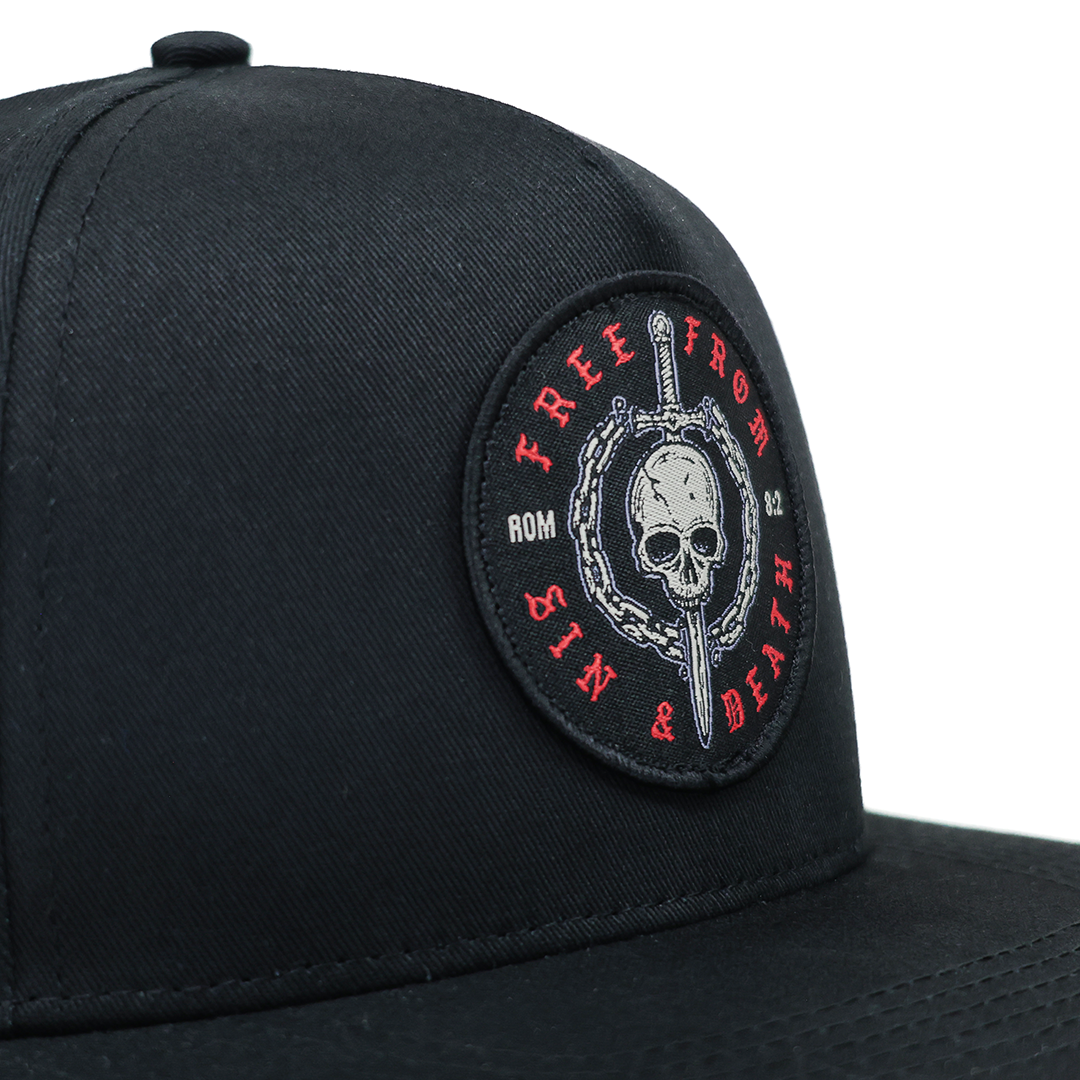 Black Christian cap with a skull and crossbones emblem on a white background. Design inspired by Romans 5:8.