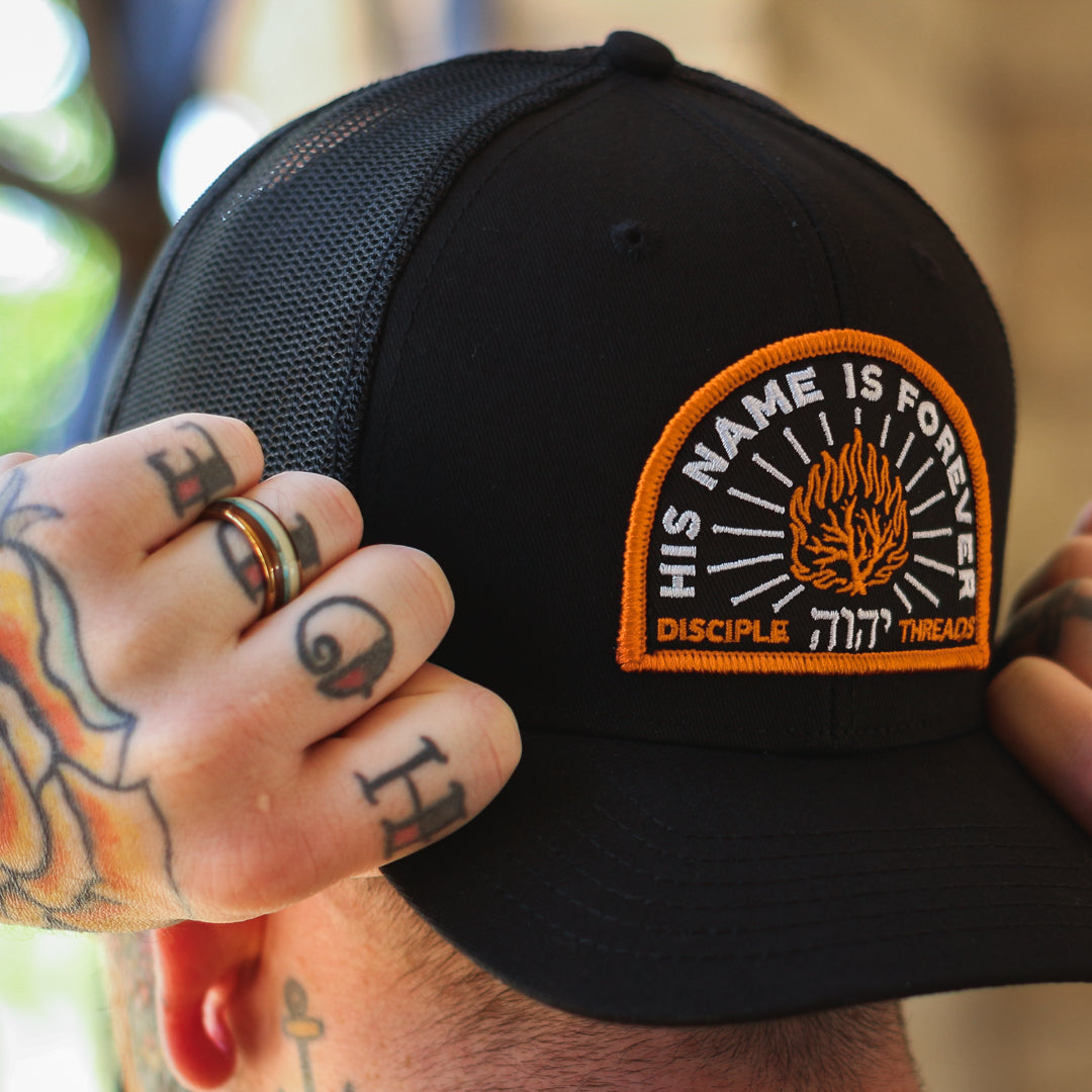 Black cap with a patch reading 'His Name is Forever' held by a person with tattoos.