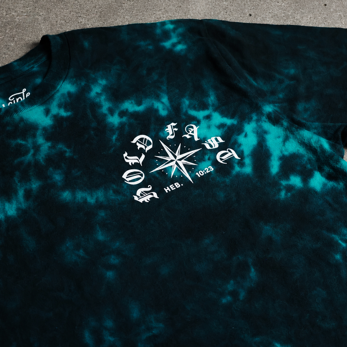 Flat lay photo of a teal and black crystal wash shirt. Hebrews 10:23 design with a stormy compass rose illustration.