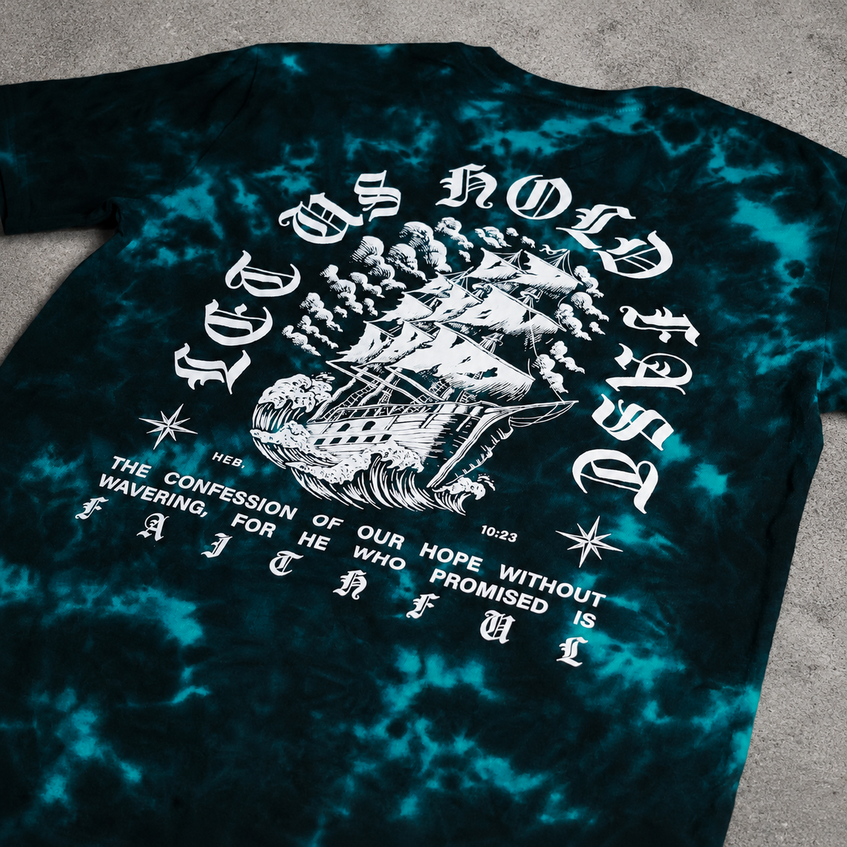 Flat lay photo of a teal and black crystal wash shirt. Hebrews 10:23 design with a stormy ship illustration.