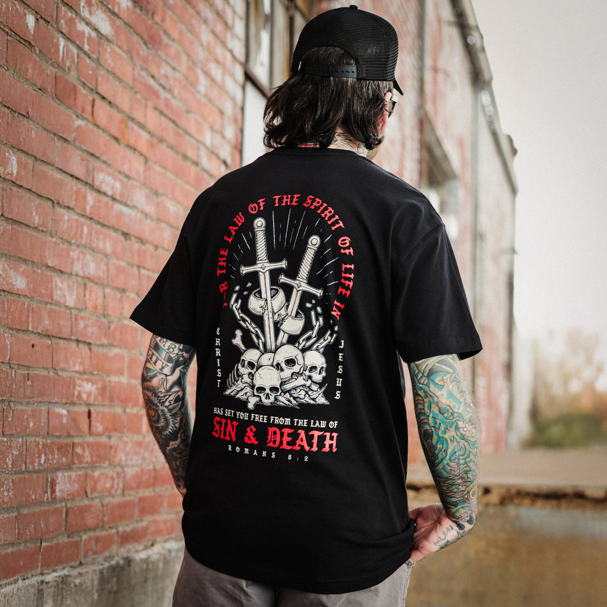 Person wearing a black, Christian t-shirt with a graphic design and text, standing against a brick wall.