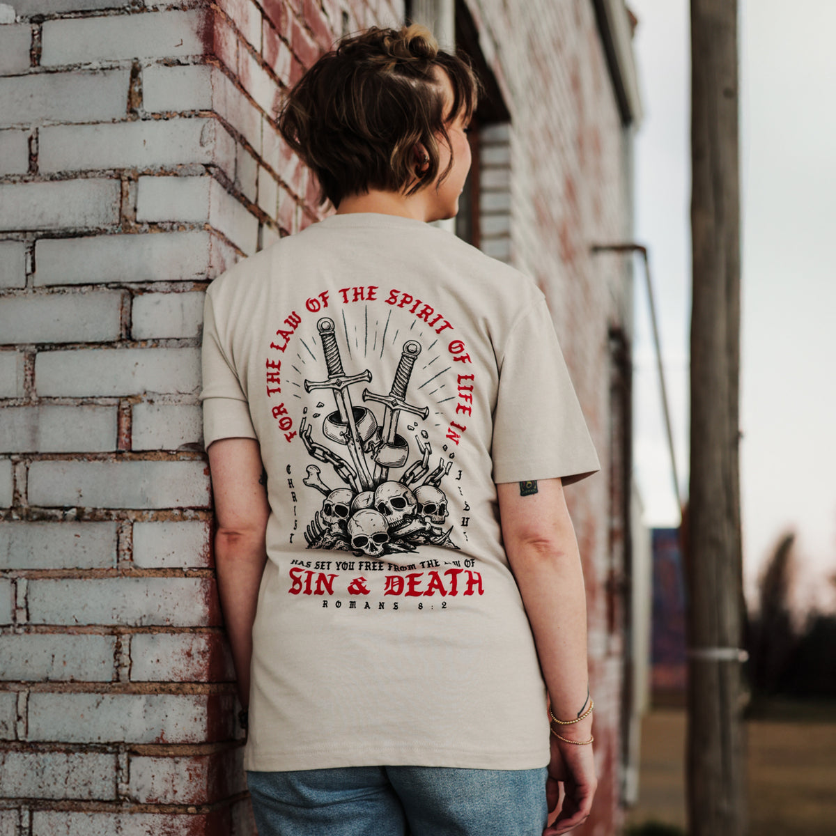 Person wearing a t-shirt with a graphic design and text, leaning against a brick wall.