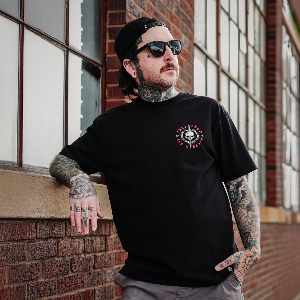 Man wearing a black t-shirt with a faith based logo, leaning against a brick wall.