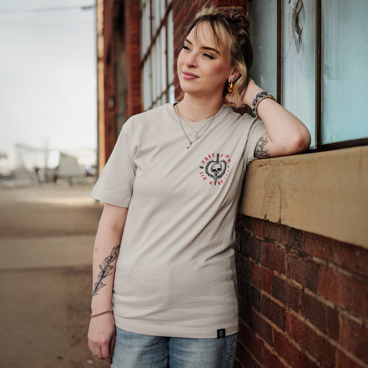 Woman wearing a beige t-shirt with a Christian design, leaning against a brick wall.