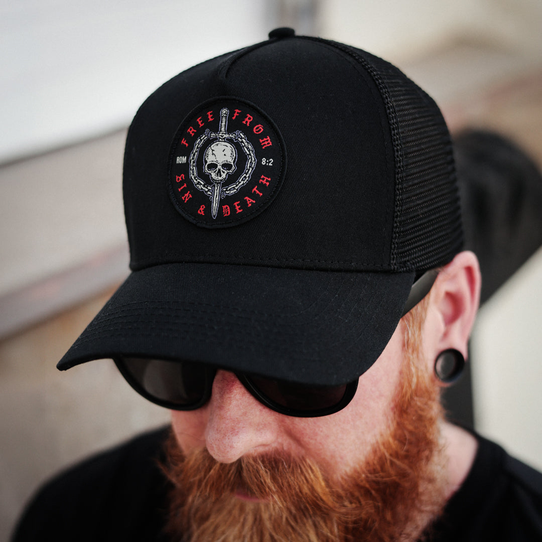 Black Christian trucker cap with a skull and sword emblem on a white background. Design inspired by Romans 5:8.