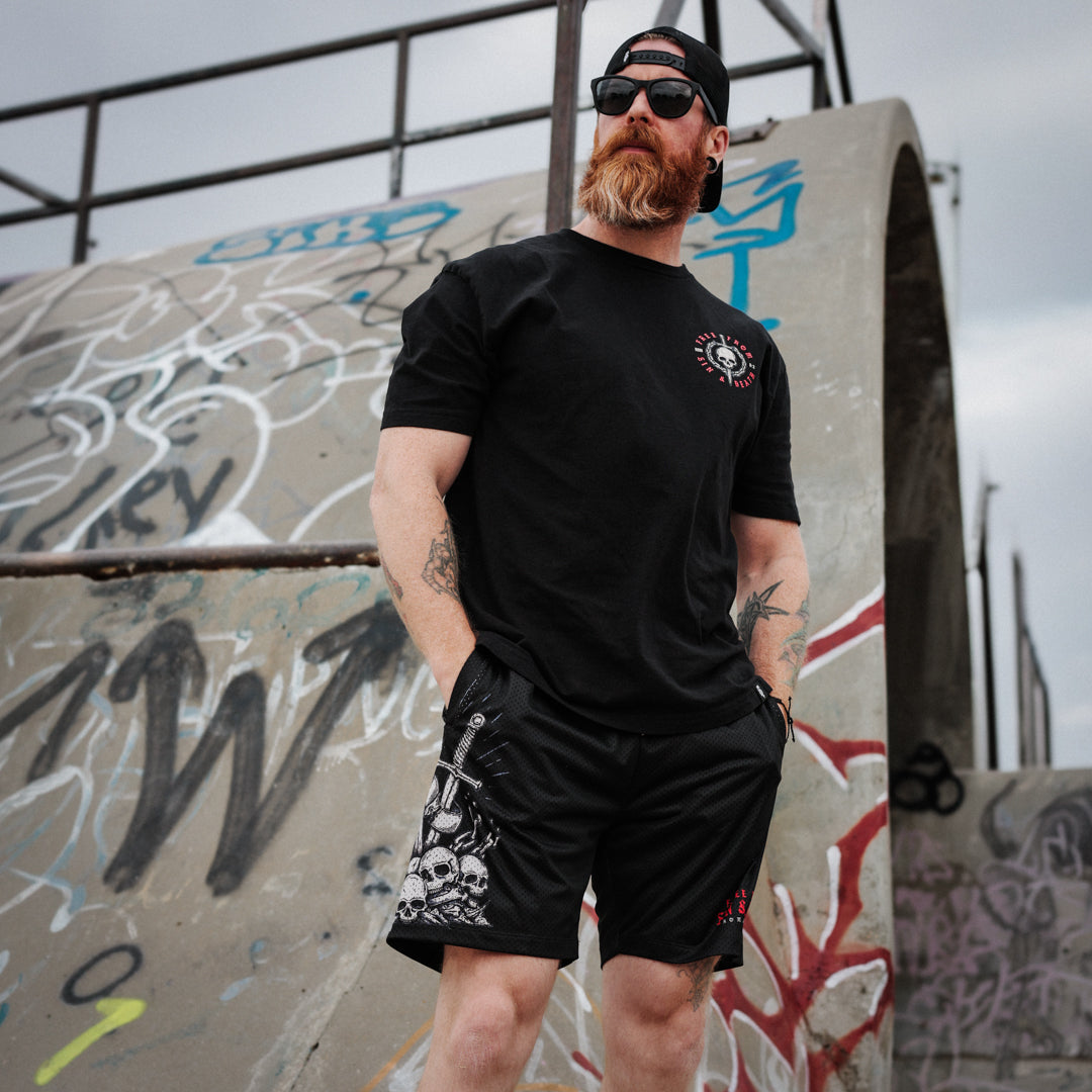 Man wearing a black Christian t-shirt and shorts with tattoos, standing in front of a graffiti-covered wall.