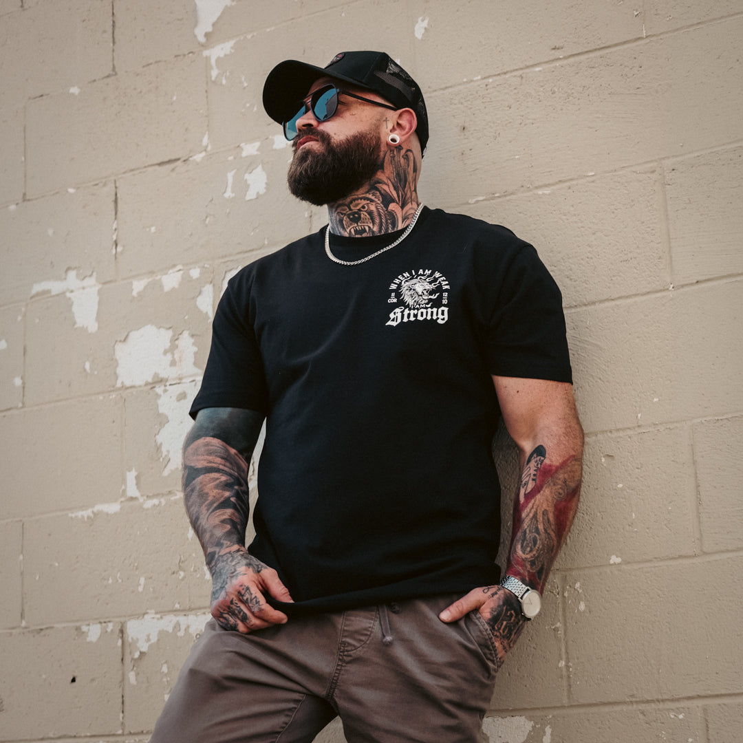Man with tattoos wearing a black Christian t-shirt with a lion and text 
standing against a textured wall.