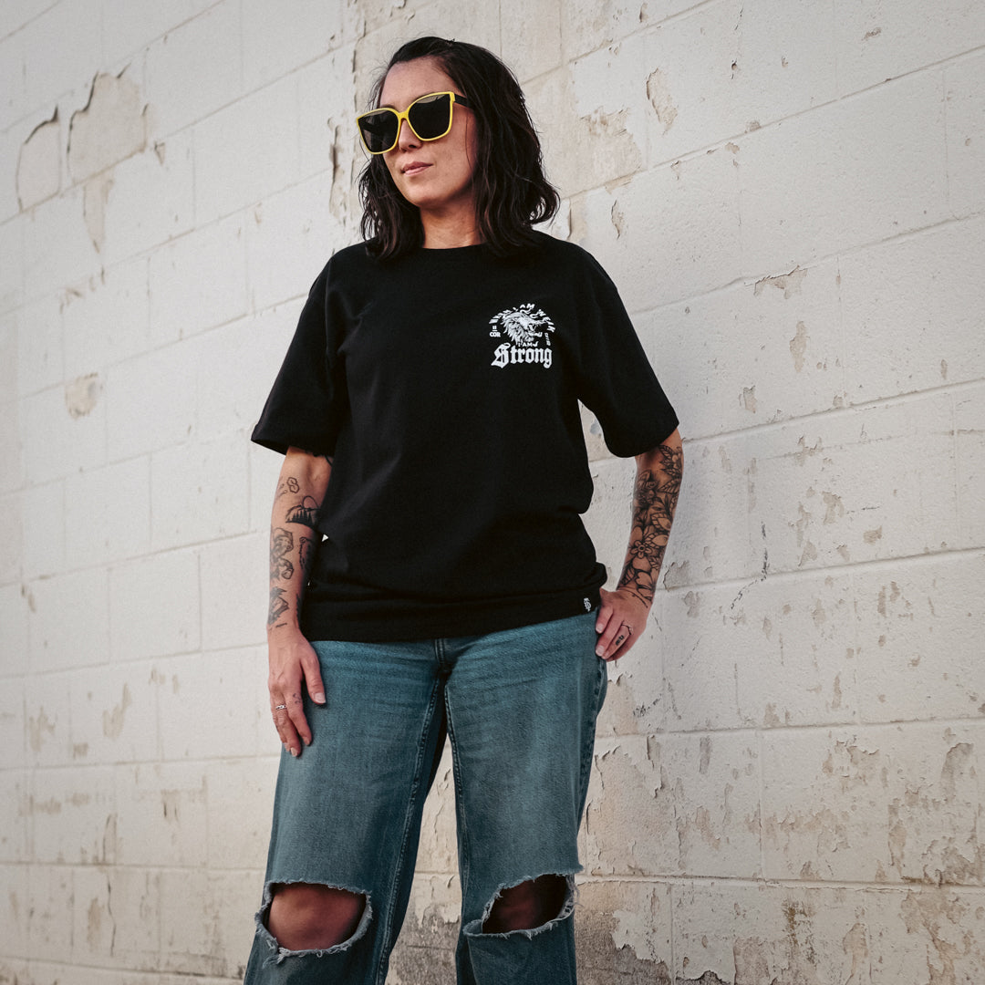 Woman wearing a black t-shirt with text and graphics, blue jeans, and sunglasses against a textured white wall.