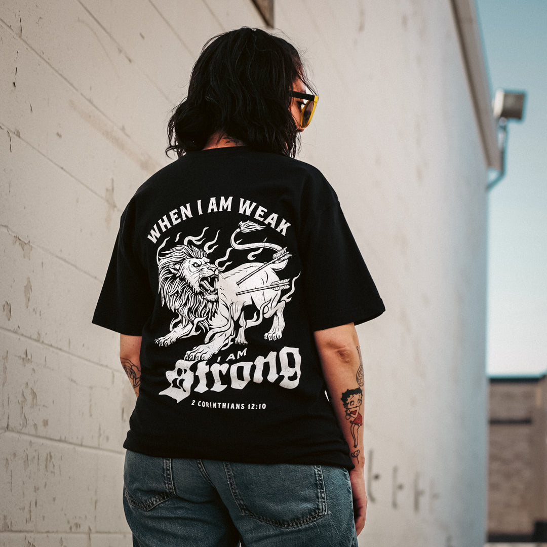Person wearing a black t-shirt with a graphic design and text on the back, standing against a light-colored wall.