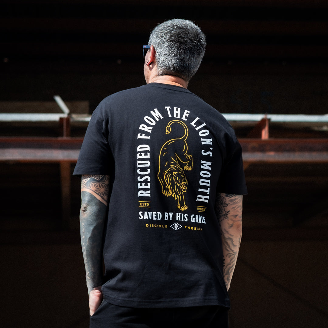 A man viewed from behind wearing a black t-shirt with a large yellow graphic of a lion and the text 'RESCUED FROM THE LION'S MOUTH SAVED BY HIS GRACE' on the back, standing in a dark industrial setting.