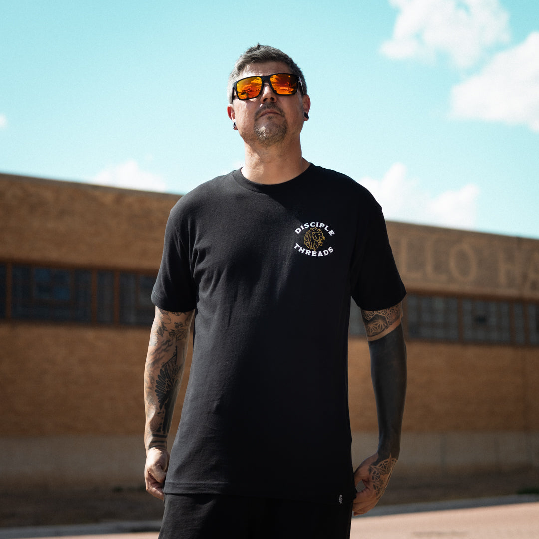 A man wearing a black crewneck t-shirt with a white circular text on the left chest that reads 'DISCIPLE THREADS' along with a lion head illustration, paired with orange-tinted sunglasses, standing outdoors in front of a brick building under a blue sky.