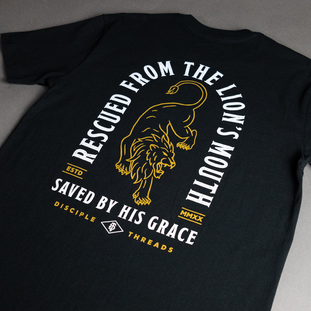 A black short-sleeve t-shirt laid flat on a gray surface, showing the back side with a large graphic print. The design features a golden lion in the center, surrounded by white and gold text reading 'RESCUED FROM THE LION'S MOUTH' and 'SAVED BY HIS GRACE', with 'DISCIPLE THREADS' at the bottom.