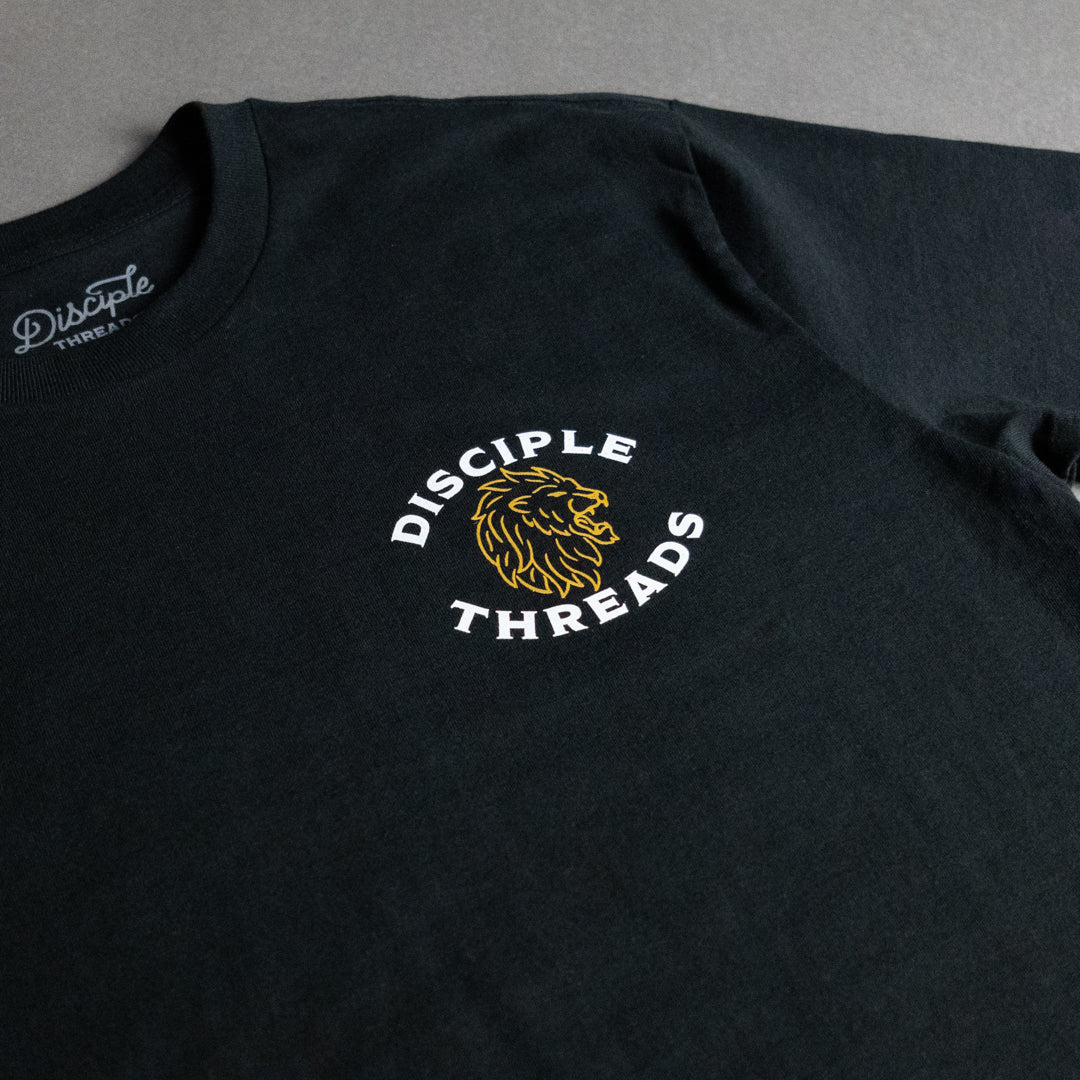 A close-up of a black t-shirt featuring a circular graphic on the chest with a yellow-gold lion head and the words 'DISCIPLE THREADS' in white, with the brand name also printed inside the collar.