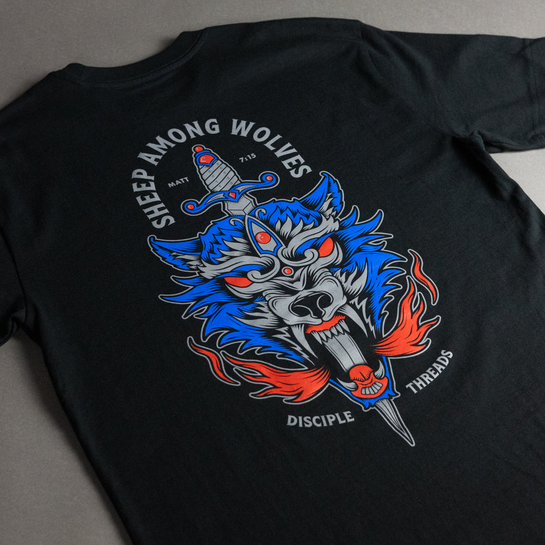 A black short-sleeve t-shirt laid flat on a gray surface, featuring a large graphic print on the back with a blue wolf head, red eyes, a dagger, flames, and the text 'SHEEP AMONG WOLVES', 'MATT 7:15', 'DISCIPLE THREADS'.