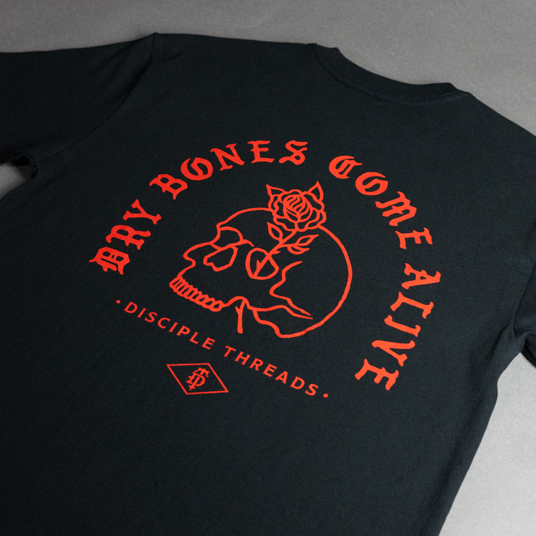A black t-shirt laid flat on a gray surface, featuring a prominent orange-red graphic on the back with the text 'DRY BONES GOME ALIVE', a skull with a rose, and 'DISCIPLE THREADS'.