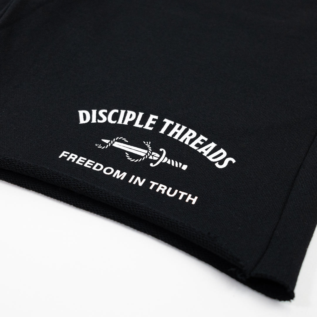 Close-up of a black fleece sweat shorts, featuring a white screen printed graphic that reads 'DISCIPLE THREADS' and 'FREEDOM IN TRUTH' with a dagger and rope motif.
