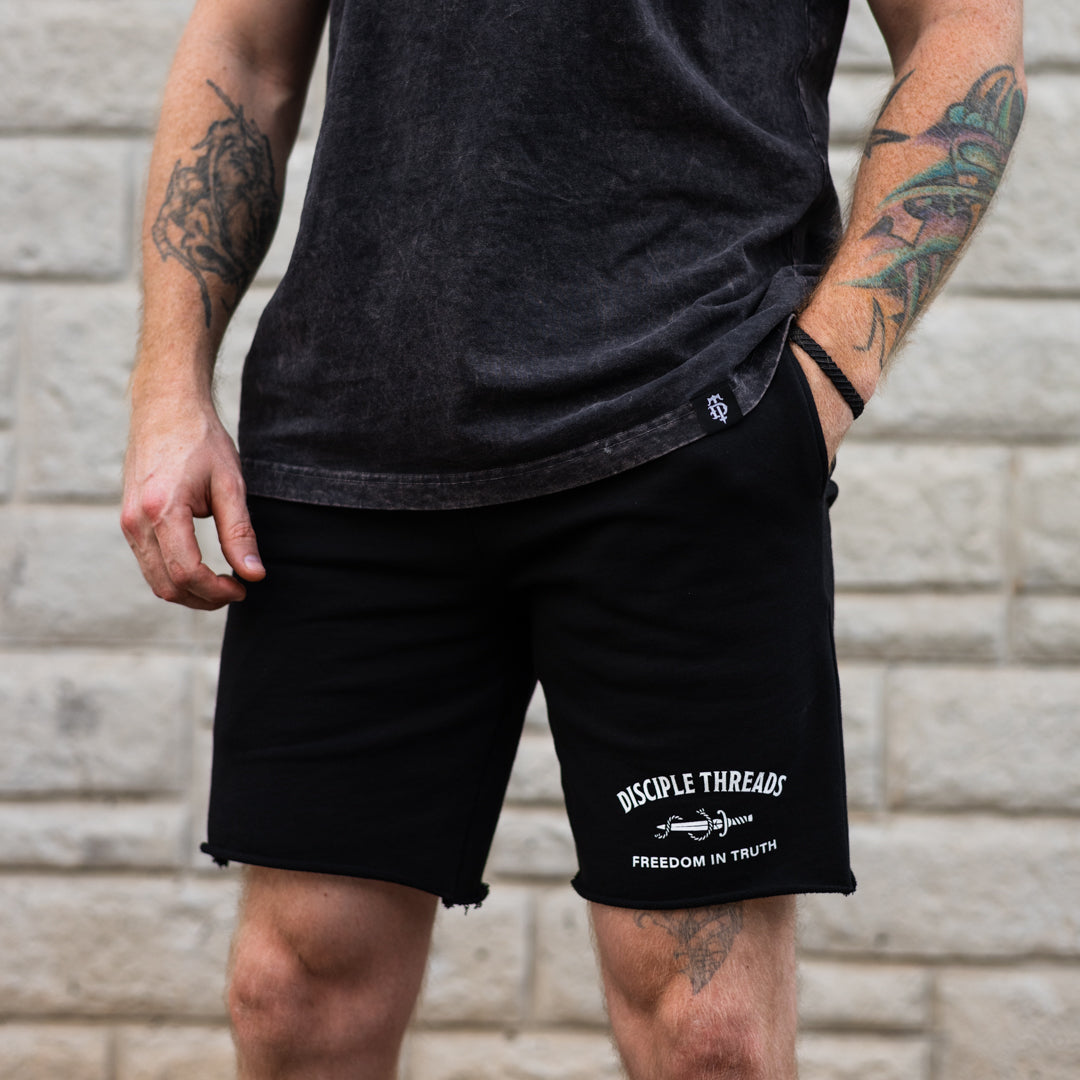 A man wearing black athletic shorts with white printed text reading 'DISCIPLE THREADS' and 'FREEDOM IN TRUTH' on the left leg, paired with a dark charcoal t-shirt, standing against a light gray brick wall.