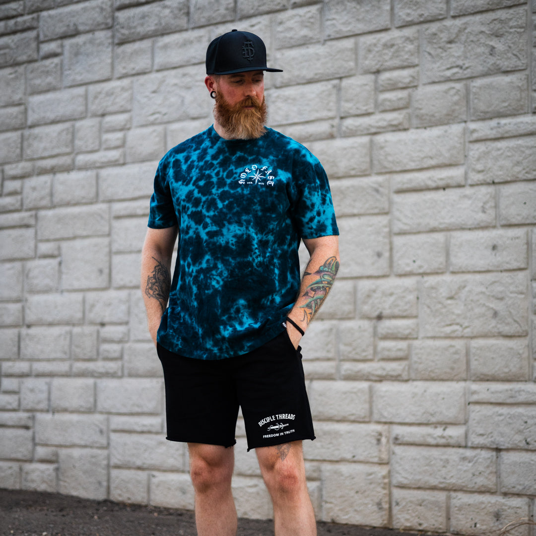 A man with a red beard and tattoos stands against a gray brick wall wearing a blue and black tie-dye t-shirt with a white chest logo, black shorts, and a black baseball cap.