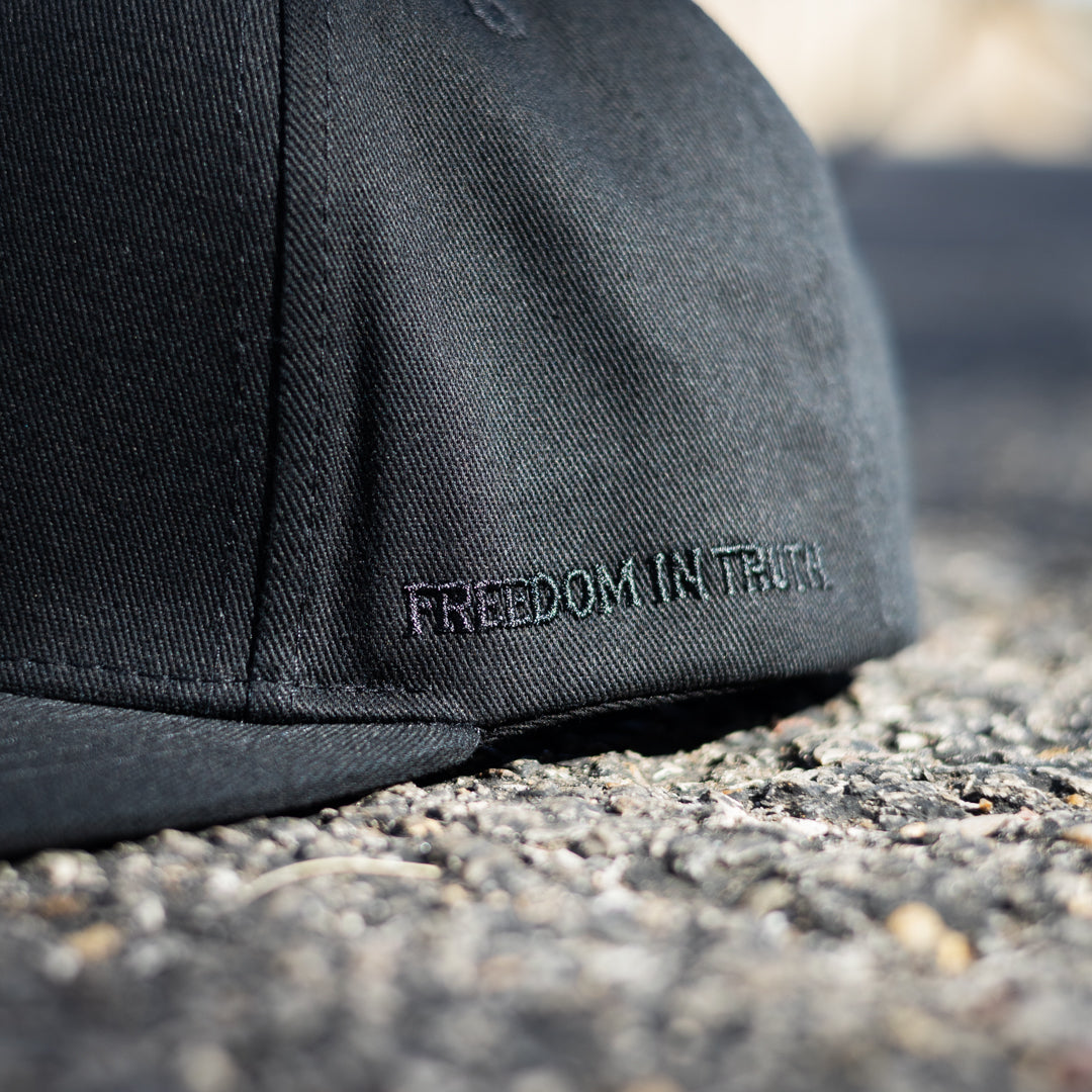 Close up of a black flat bill hat with "Freedom in Truth" embroidered on the side.
