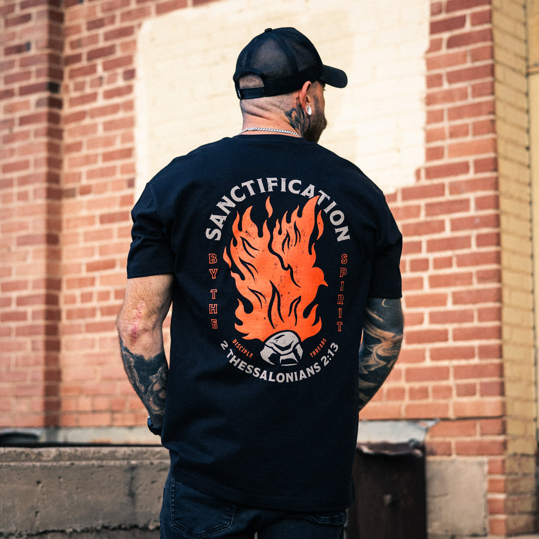 Male model wearing the black Sanctification T-shirt by a brick wall, showing the large orange flaming dove and coal graphic with Sanctification by the Spirit and 2 Thessalonians 2:13.