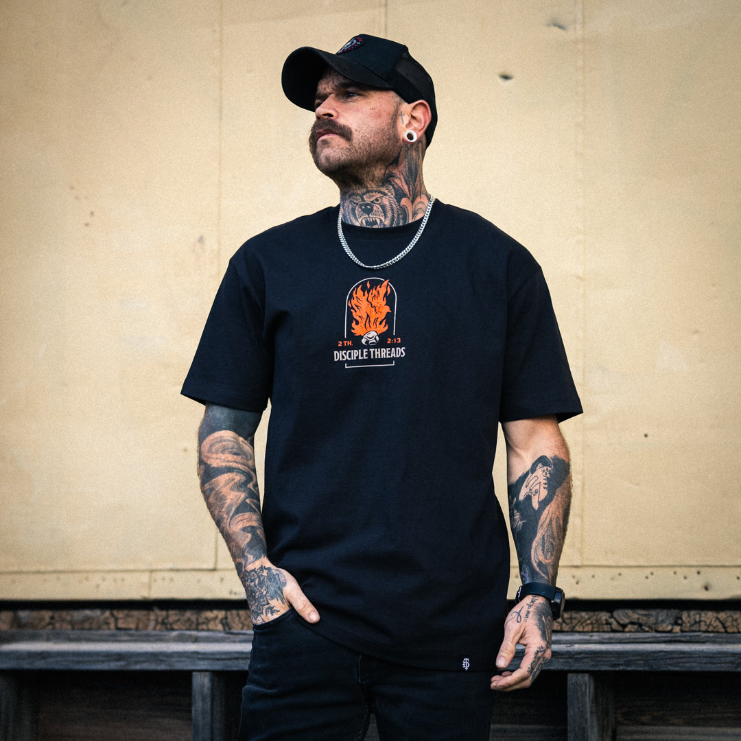 Male model wearing the black Sanctification T-shirt against a tan wall, showing the small orange flaming dove and Disciple Threads chest graphic, 2 Thessalonians 2:13.