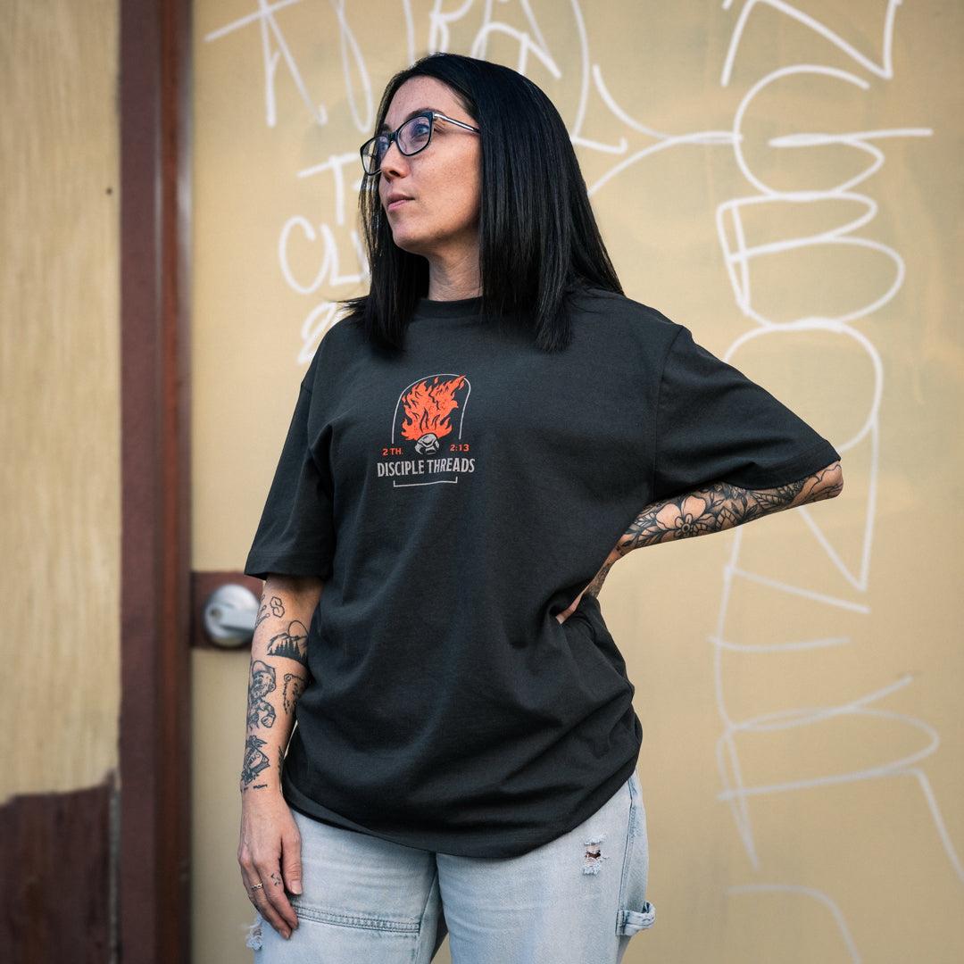 Female model wearing the dark grey Sanctification T-shirt against a graffiti wall, showing the small orange flaming dove and Disciple Threads chest graphic, 2 Thessalonians 2:13.