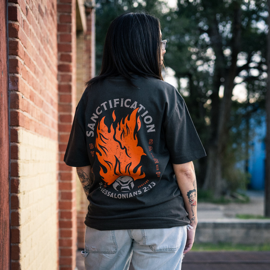 Female model wearing the dark grey Sanctification T-shirt by a brick wall, showing the large orange flaming dove and coal graphic with Sanctification by the Spirit and 2 Thessalonians 2:13.