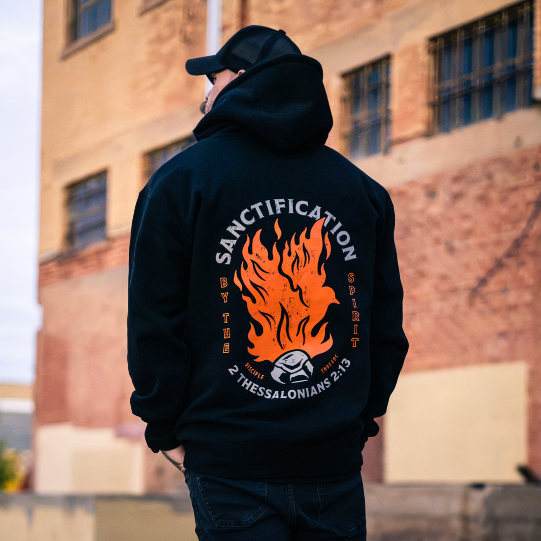 Male model wearing the black Sanctification Pullover Hoodie by a brick building, showing the large orange flaming dove and coal graphic with Sanctification by the Spirit and 2 Thessalonians 2:13.