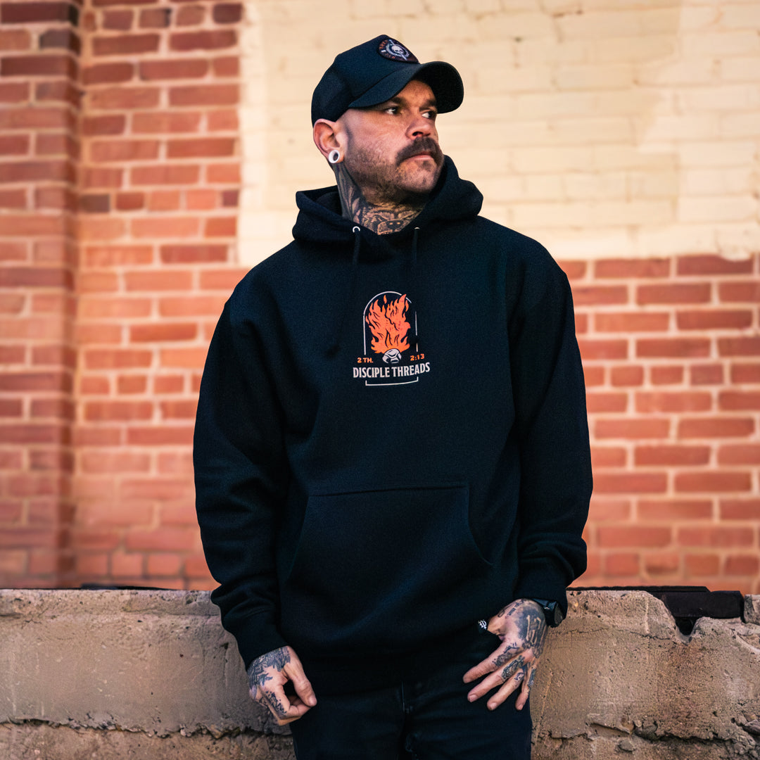 Male model wearing the black Sanctification Pullover Hoodie against a brick wall, showing the small orange flaming dove and Disciple Threads chest graphic, 2 Thessalonians 2:13.