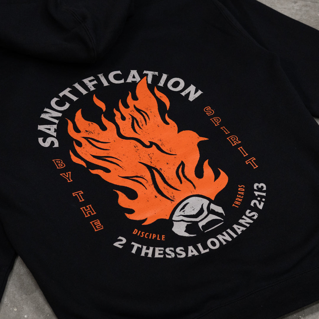 Close-up of the black Sanctification Pullover Hoodie graphic, an orange flaming dove rising from a burning coal, framed by Sanctification by the Spirit and 2 Thessalonians 2:13.
