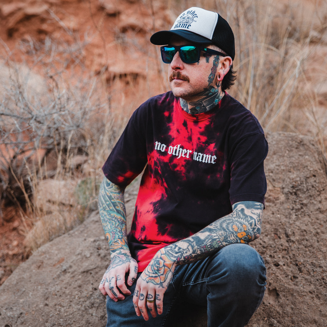 Man with tattoos wearing a black and red crystal wash shirt with 'no other name' text, sitting outdoors.