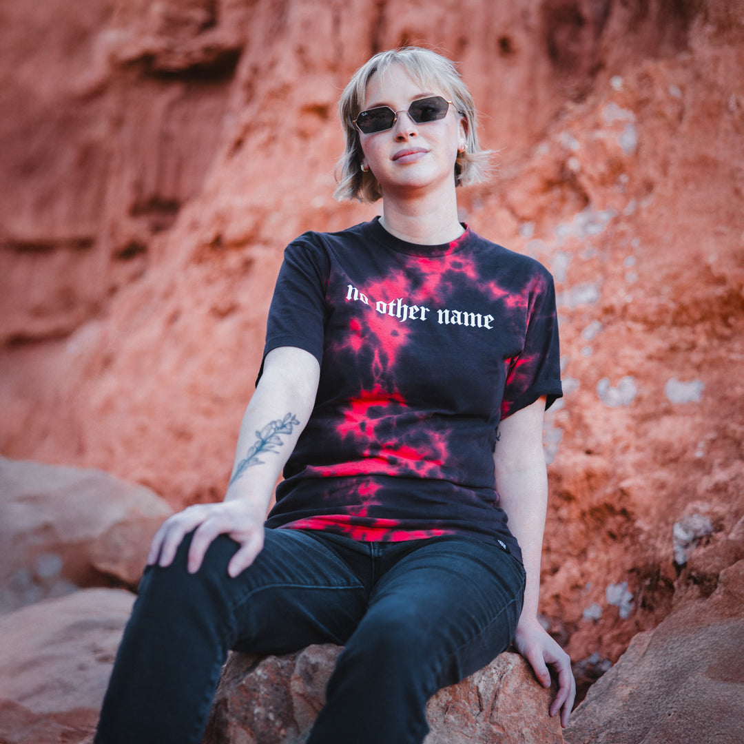Person wearing a black t-shirt with red design and text, sitting on rocks against a red rock formation.