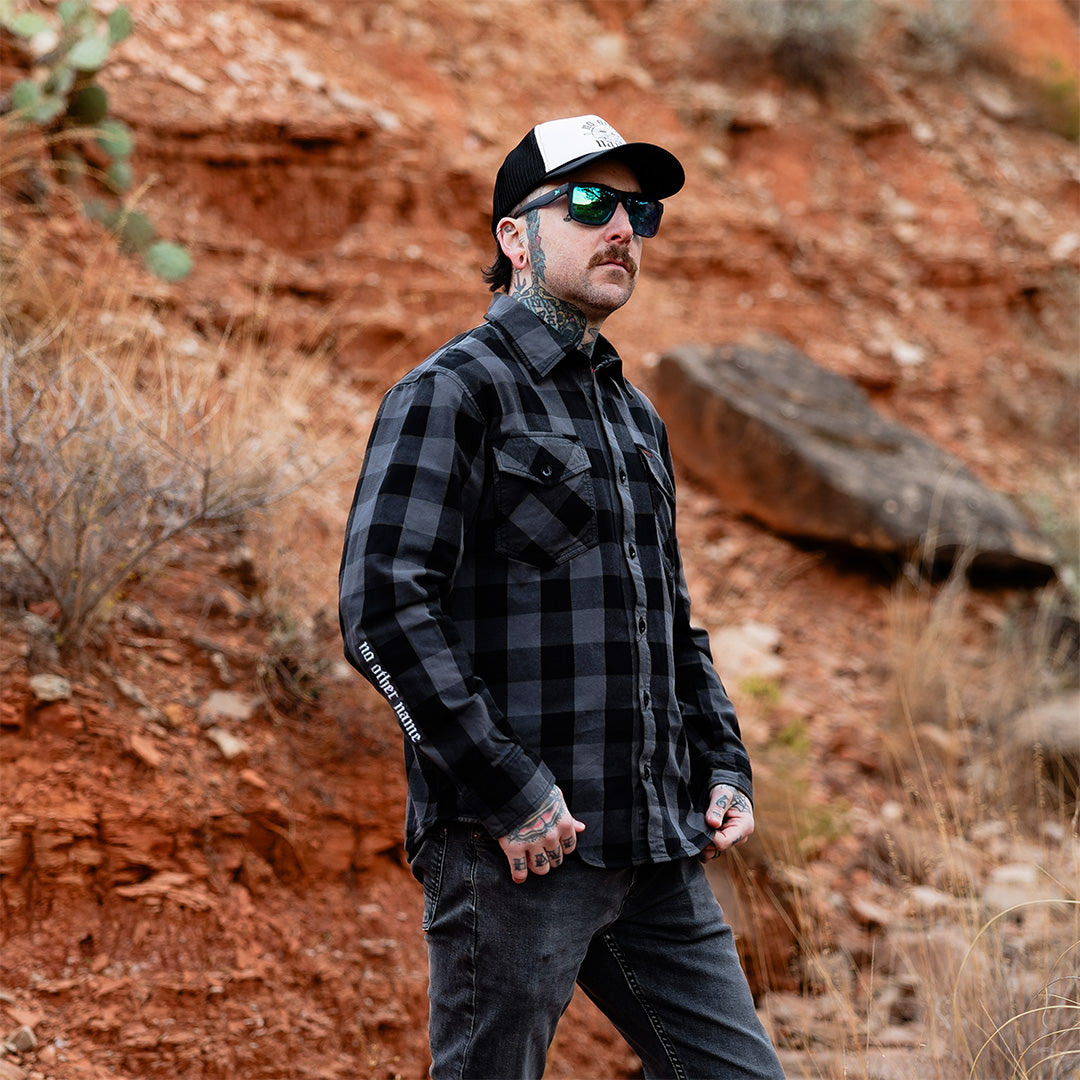 Man wearing a black and gray plaid shirt, sunglasses, and cap standing in a desert landscape.