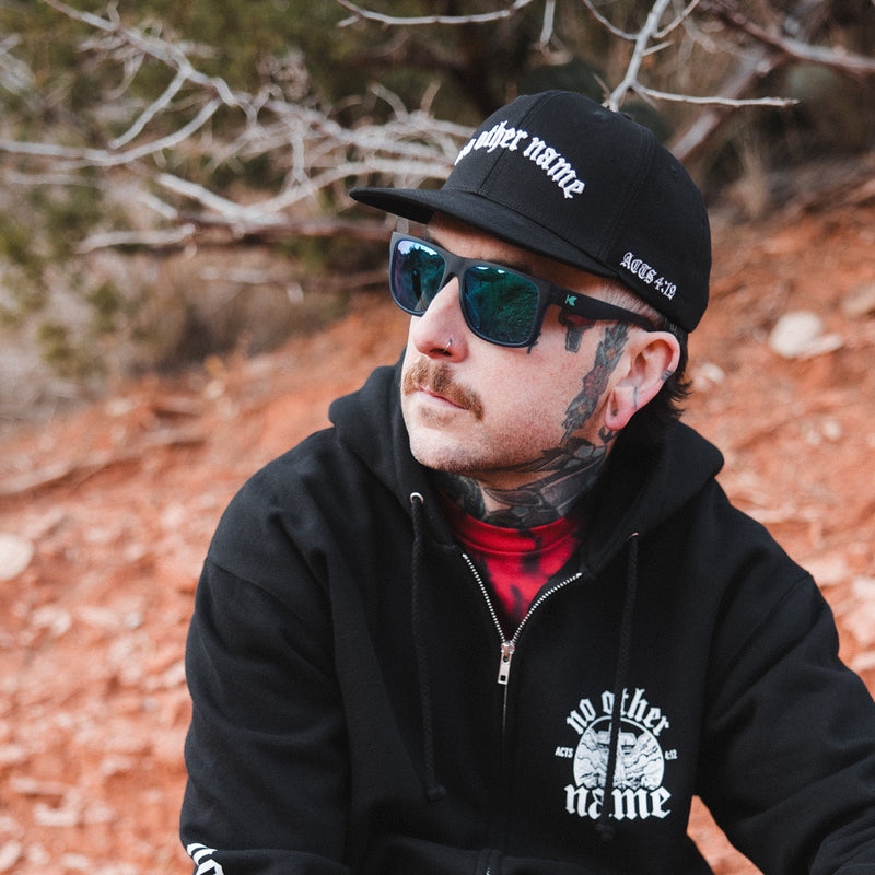 Man wearing sunglasses and a black cap with text, sitting outdoors.