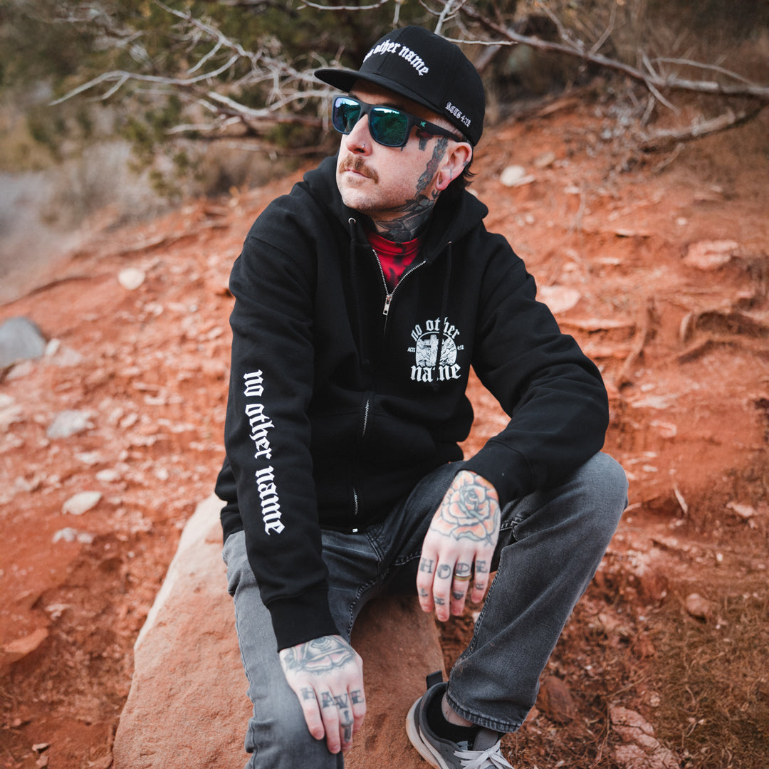 Man wearing a black hoodie with text, sunglasses, and a cap sitting on a rock in a natural setting.