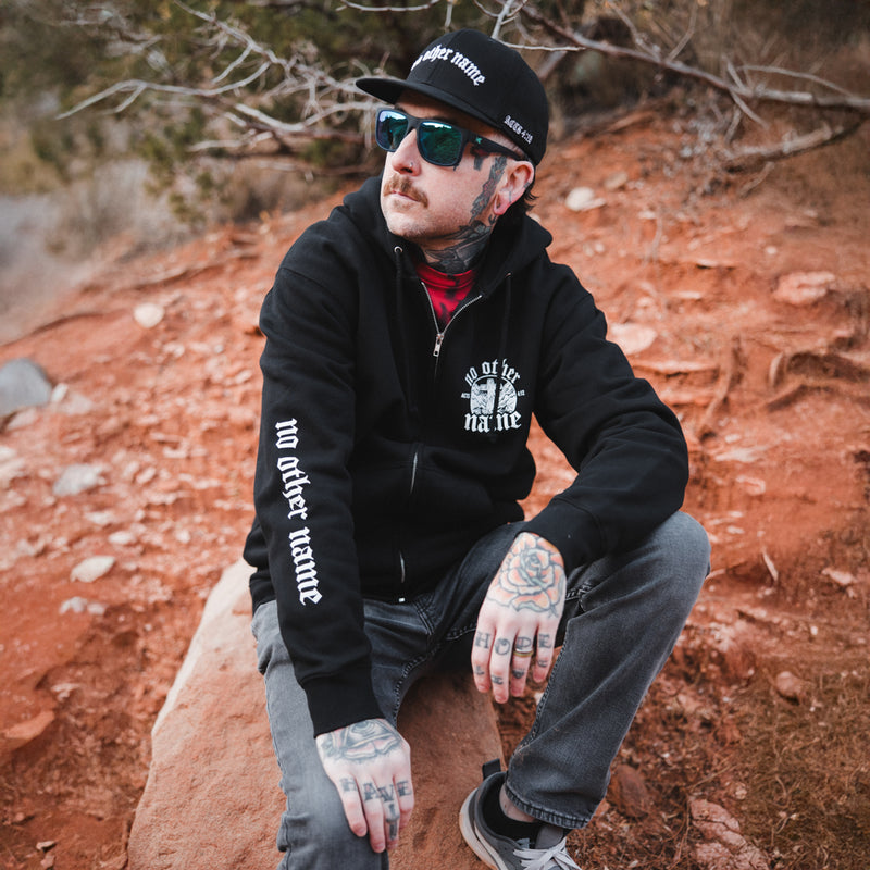 Man wearing a black hoodie with text, sunglasses, and a cap sitting on a rock in a natural setting.