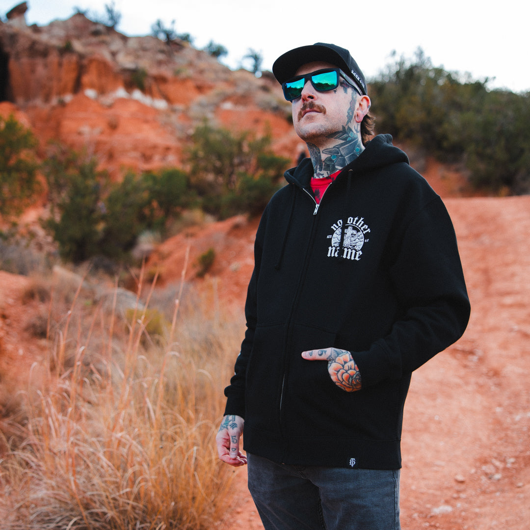 Man wearing a black hoodie with text, sunglasses, and tattoos standing in a desert landscape.