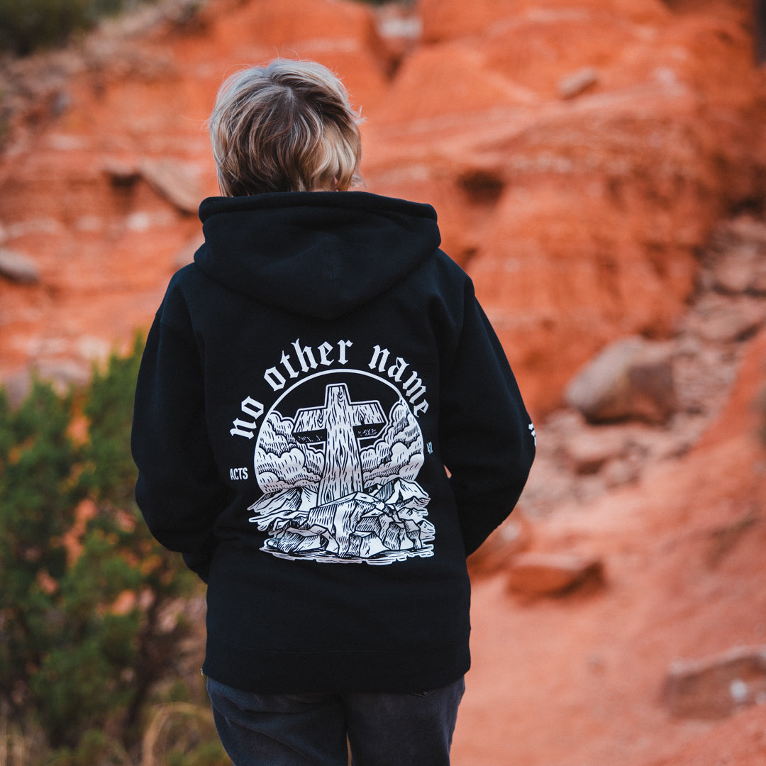 Person wearing a black hoodie with a cross design and 'no other name' text in a desert landscape.