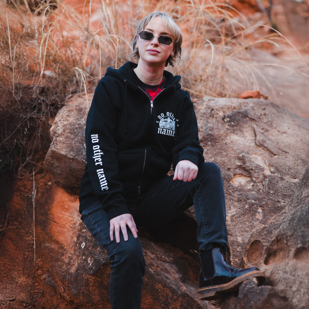 Person wearing a black hoodie with text, sitting on a rock in a natural setting