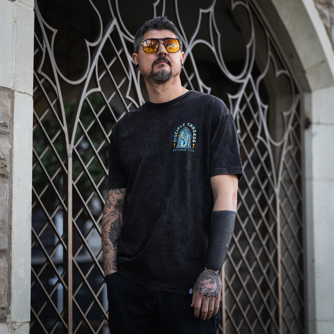 Man wearing a black t-shirt with a logo in front of an ornate metal gate.